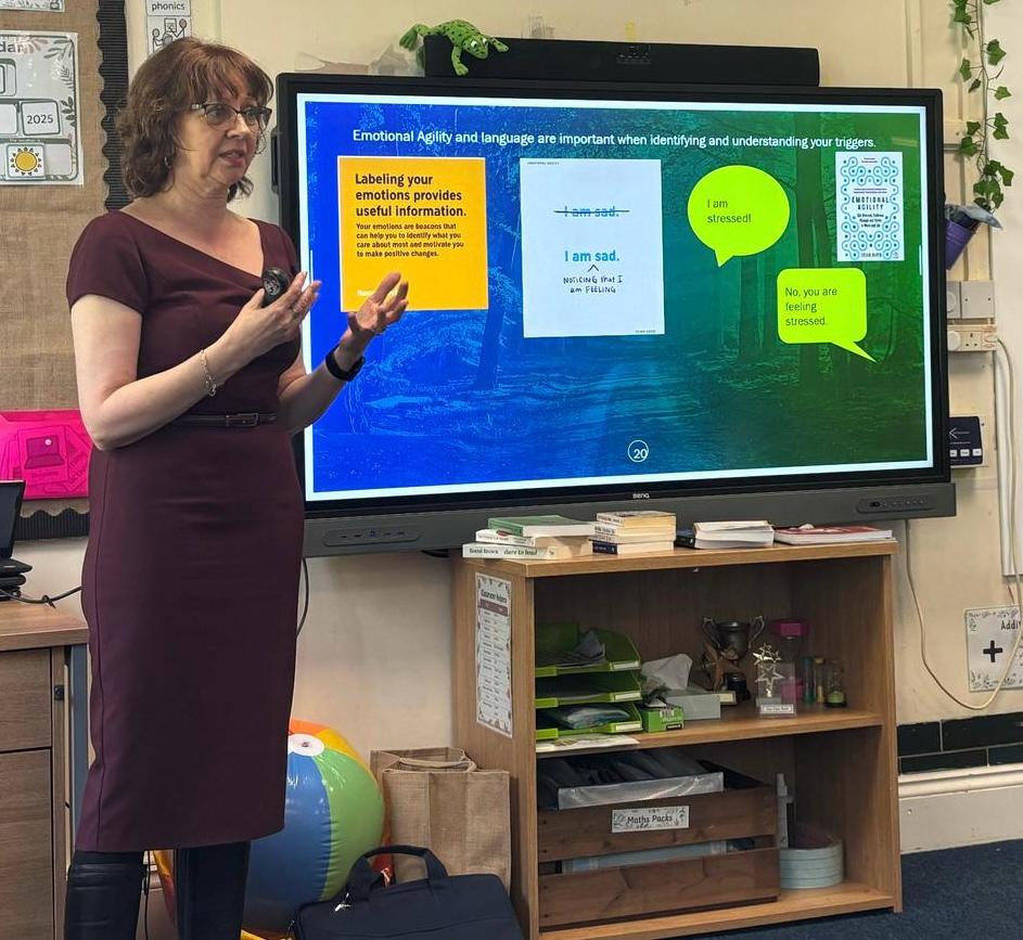 Gillian standing at the front of a classroom talking to course members with a large screen on the wall displaying course information